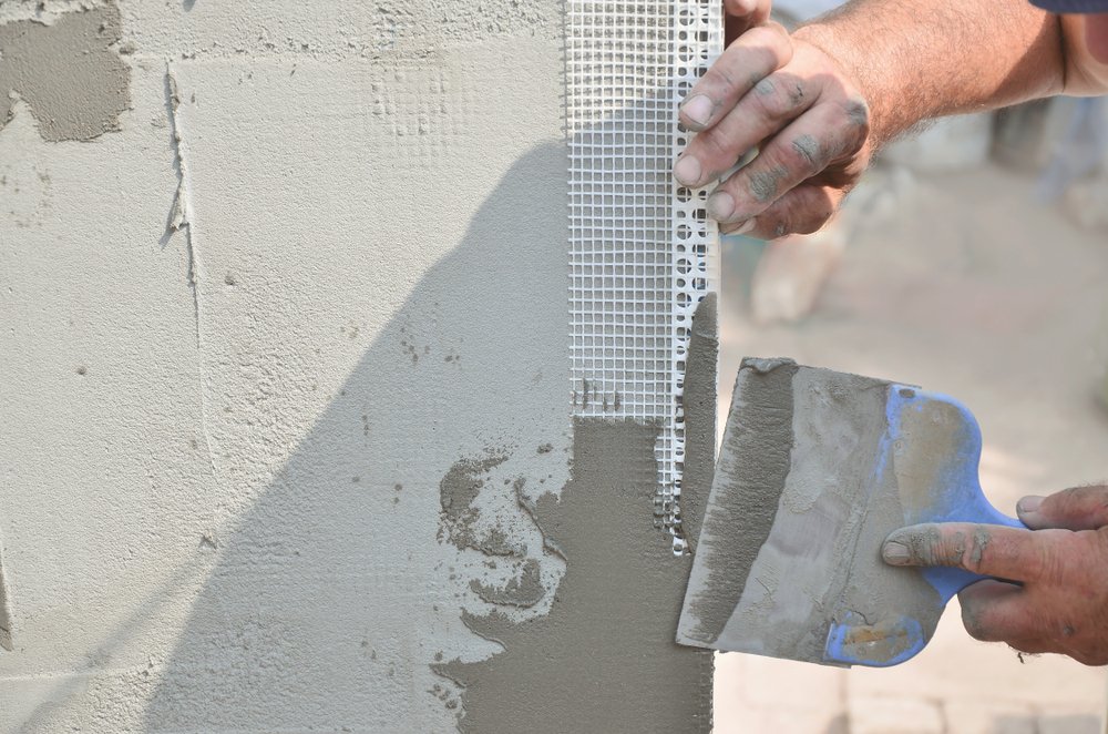 Een metalen hoekprofiel wordt geïnstalleerd op een hoek van een muur, met pleistermateriaal en een pleisterspaan op de voorgrond, terwijl een persoon het hoekprofiel stevig op zijn plaats drukt.