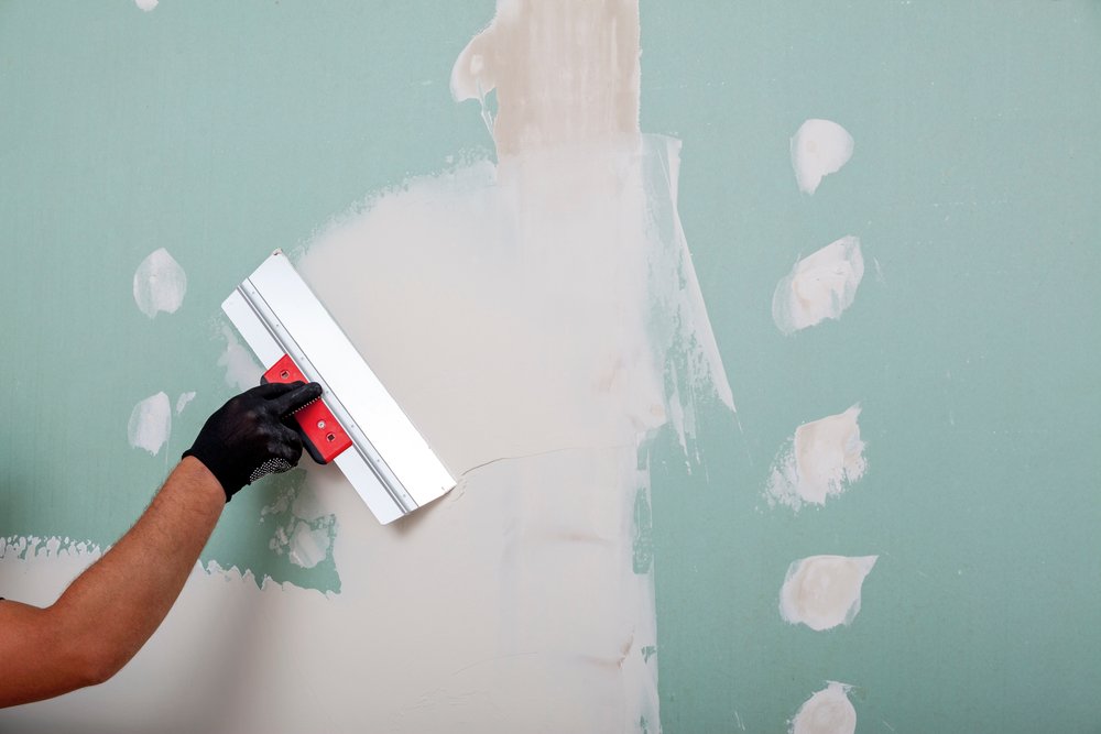 Een persoon brengt vulmiddel aan op een muur met een plamuurmes, terwijl een andere persoon de muur schuurt voor een gladde afwerking, met schuurpapier en primer op de voorgrond.