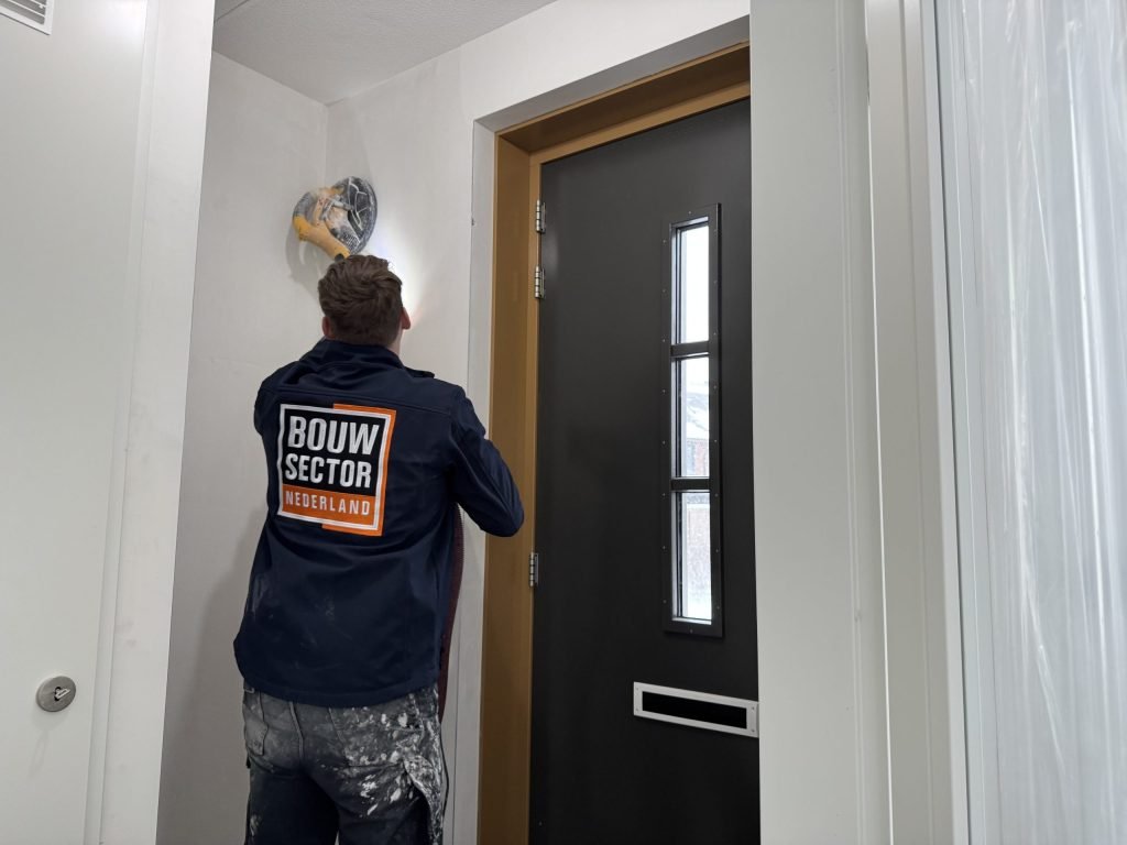 Vakman van Bouwsector Nederland in werkjas schuurt een wand boven een voordeur met een schuurmachine