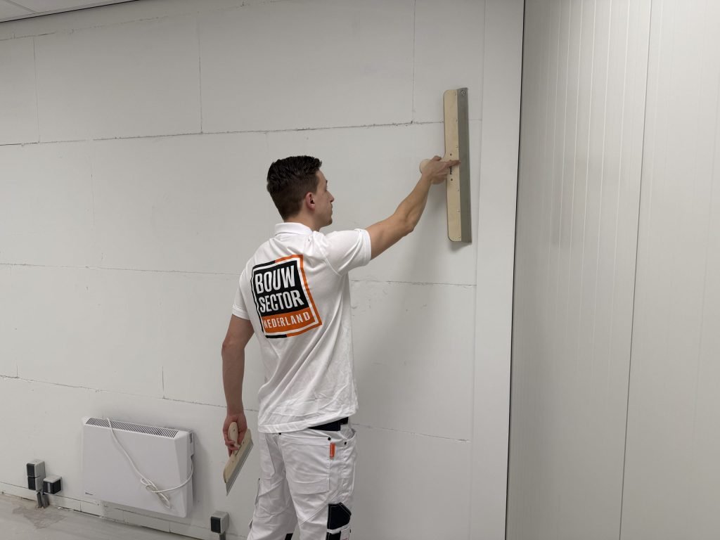 Stukadoor van Bouwsector Nederland zet een brede spaan hoog tegen een kale wand, met een tweede spaan in zijn andere hand