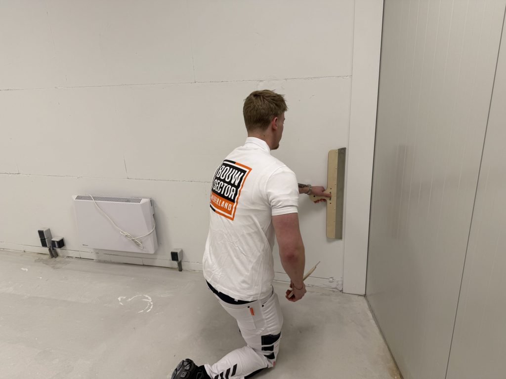 Stukadoor van Bouwsector Nederland werkt geknield met een brede spaan het onderste deel van een wand bij