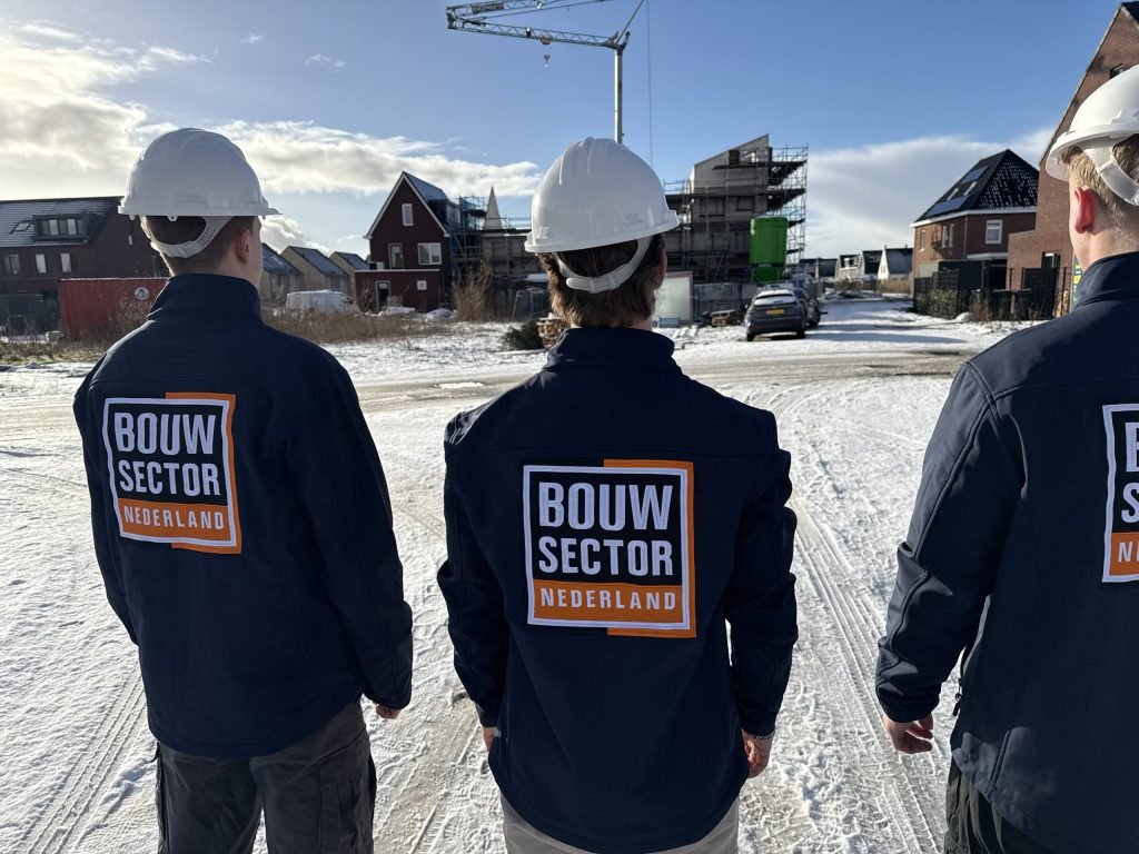 Drie vakmensen in blauwe jassen staan naast elkaar met een hijskraan boven de bouwplaats